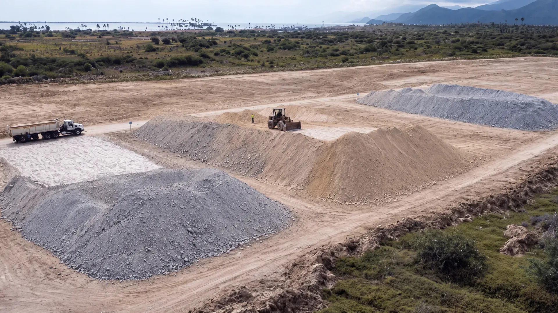 Aerial view of a stockpile yard with graded piles of mineral, a loader and a truck, with the coast and mountains beyond.