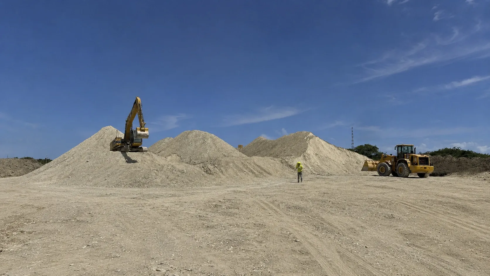 Stockyard with cones of pale clay, an excavator on top of a pile and a wheel loader alongside, under a clear blue sky.