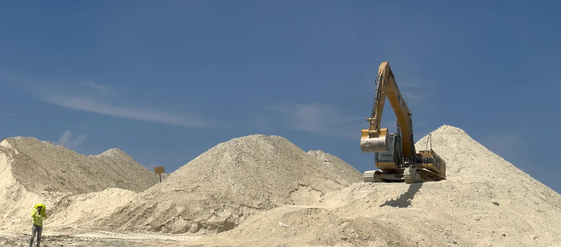 Excavator on top of stockpiles of white clay, against a clear blue sky.