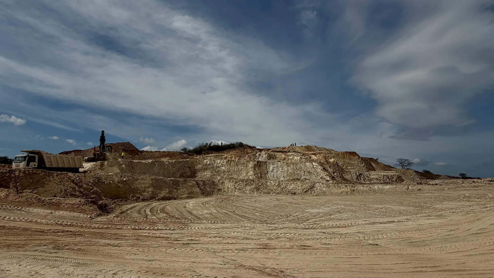 Terraced face of pale clay under a wide cloudy sky, with a haul truck and an excavator on the upper bench.