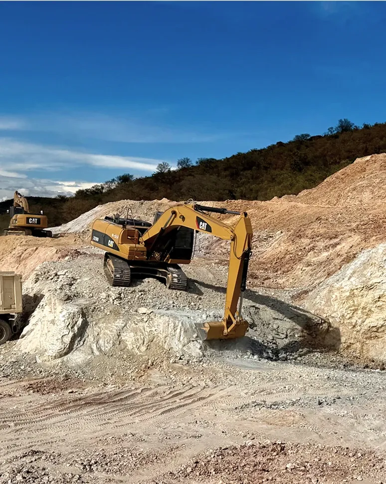 Excavator working a face of white clay, with a second machine on the bench above and hills behind.