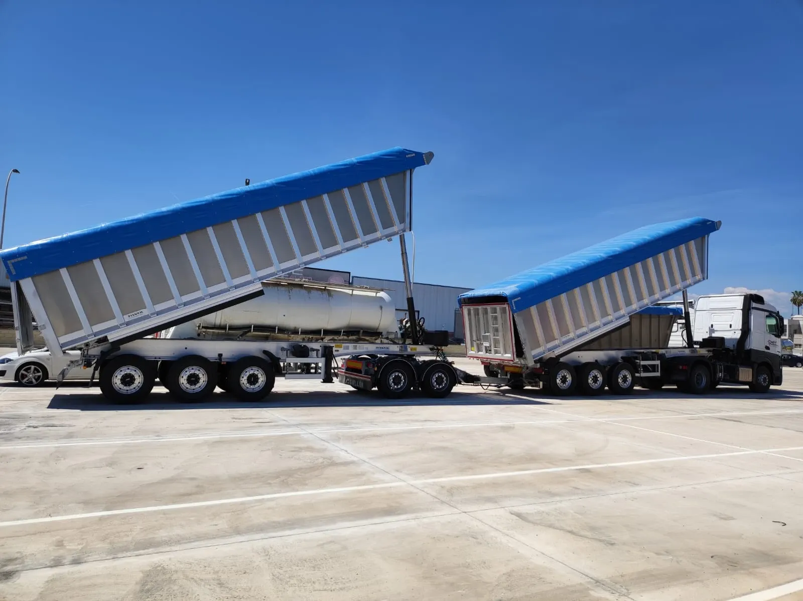 Two aluminium tipper trailers with blue covers and their bodies raised, parked on a concrete yard under a clear sky.