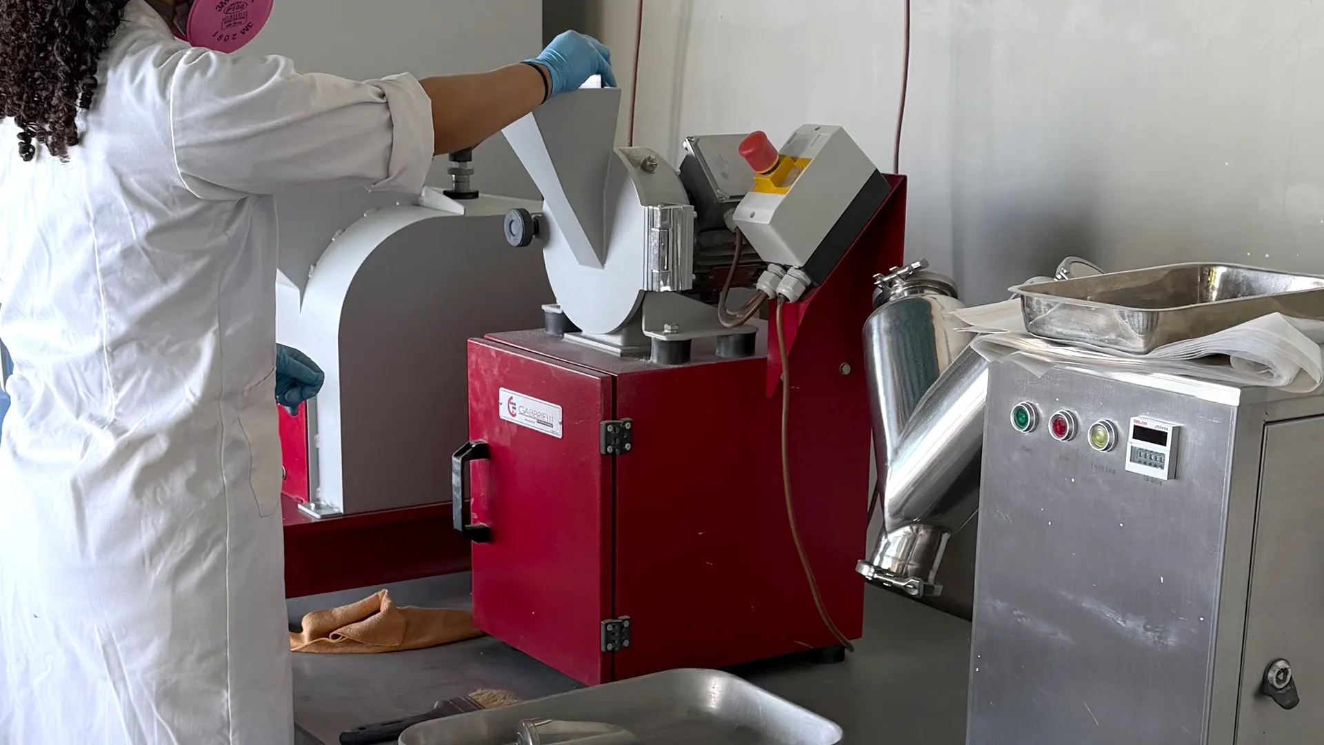 Laboratory technician in a coat and mask feeding a sample into a crusher, with a blender alongside.