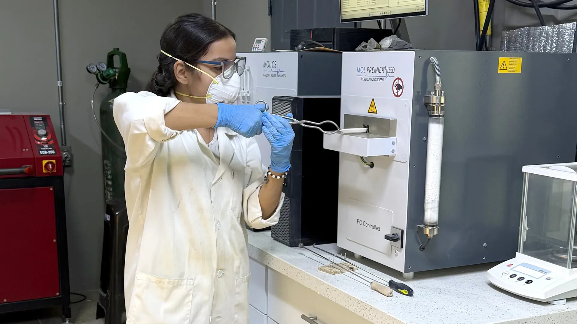 Laboratory technician in a coat and mask operating an analysis instrument at the bench.