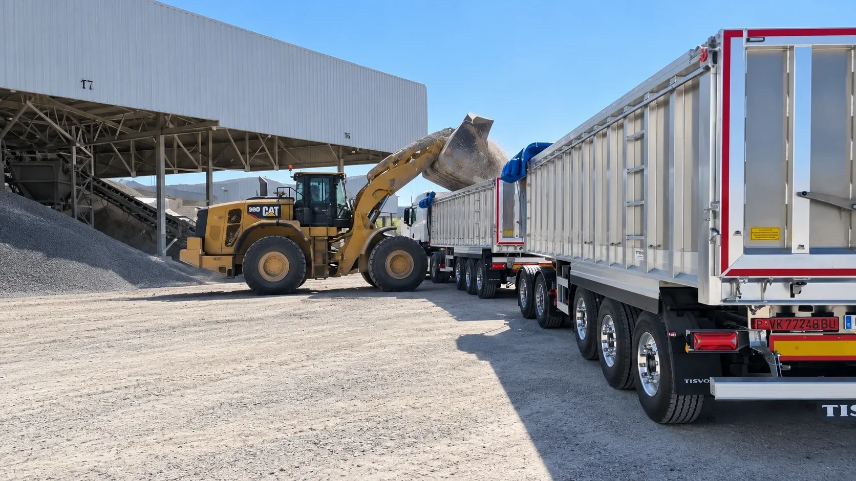 Wheel loader tipping clay into an aluminium trailer beside the covered bays of a processing plant, under a clear sky.