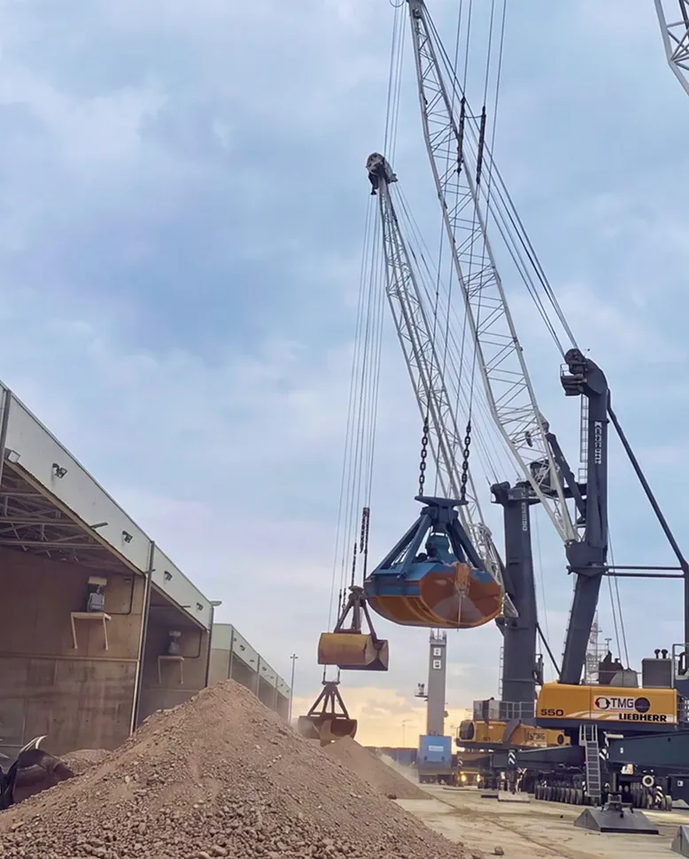 Harbour crane lowering a grab over a stockpile of mineral at the quay, with the warehouse alongside.